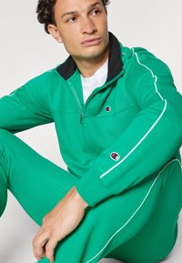 Green tracksuit made of soft fabric, featuring a black collar, white piping details, and a logo on the chest and sleeve. Mid-zip design.