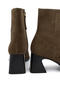 Botas de tobillo de ante marrón con cremallera, que cuentan con un distintivo tacón grueso negro y una textura suave. El diseño incluye una silueta elegante.