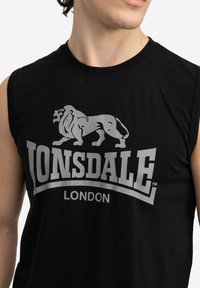 Juodas be rankovių marškinėlius su dideliu sidabriniu liūto logotipu ir įspūdinga "LONSDALE LONDON" užrašu, spausdintu ant priekinės pusės.