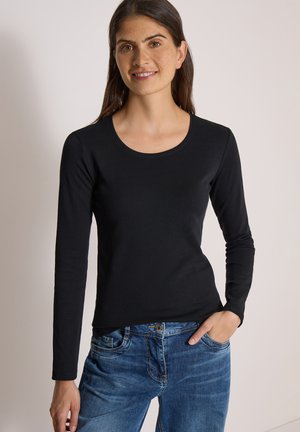 Femme aux longs cheveux foncés portant un t-shirt noir à manches longues et un jean bleu, debout avec une main dans la poche, souriant légèrement.