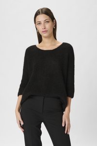 Melnā adīta džemperis ar brīvu piegriezumu, plašu laivu veida kakla izgriezumu un trīs ceturtdaļu piedurknēm, apvienots ar tumšām biksēm. Mīksta tekstūra ar nedaudz pūkainu izskatu.