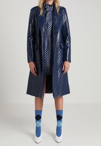 Cappotto lungo in ecopelle trapuntato di colore blu navy, con rever, abbinato a un vestito a pois e calzini blu con motivo argyle, completato da décolleté bianchi a punta.