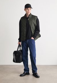 Gilet trapuntato verde sopra un maglione nero, abbinato a jeans blu e scarpe nere. Tiene in mano una borsa in pelle nera. Sfondo minimalista.