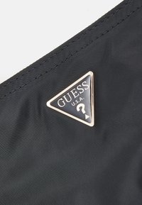 Sort nylon stof med et trekantet sølv- og sort Guess-logo mærke, der viser brandnavnet og et spørgsmålstegn design.