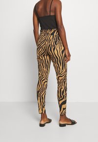 Pantalons rayés zébrés marron et noir, près du corps avec poches, associés à un débardeur noir et des sandales noires à glissière, mettant en avant une texture lisse.