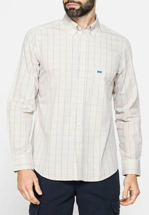 Uomo che indossa una camicia beige a quadri a maniche lunghe con bottoni e pantaloni scuri, in piedi con le mani rilassate davanti a uno sfondo semplice.