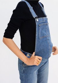 Denim overalls with adjustable straps, front pocket, and button closures. Features a relaxed fit and classic blue wash. Paired with a black top.