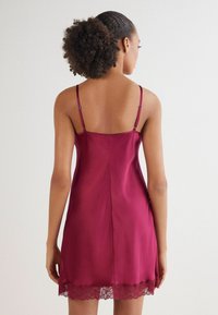 Vestido de satén burdeos tipo slip con tirantes ajustables y dobladillo de encaje. Textura suave con un silueta ajustada y un detalle de costura en la parte posterior.