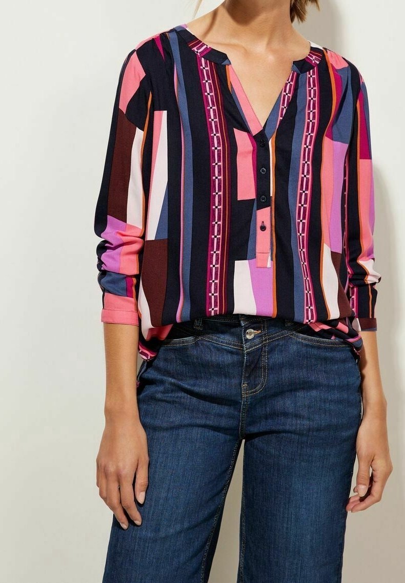 Femme portant un chemisier rayé multicolore avec des boutons et un jean bleu foncé, debout avec une main détendue le long du corps.