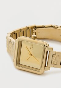 Kultainen neliönmuotoinen kello, jossa on kiiltävä näyttö, minimalistinen muotoilu ja kultainen metalliranneke, jossa on teksturoidut yksityiskohdat linkeissä.