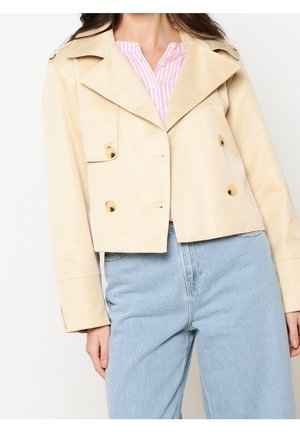 Femme portant une veste beige courte à double boutonnage avec des boutons dorés, une chemise rose rayée et un jean taille haute bleu clair.