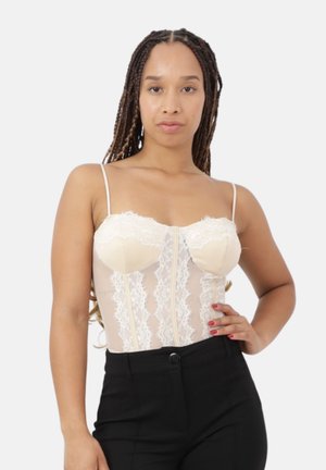 Femme aux longues tresses portant un corset beige en dentelle et un pantalon noir, posant une main sur la hanche devant un fond uni.