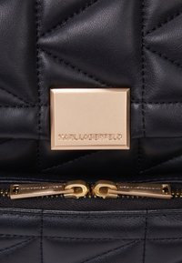 Czarna pikowana skórzana torba z metalową złotą tabliczką z logo oraz podwójnymi złotymi suwakami z grawerowanym wzorem. Widoczna faktura powierzchni.