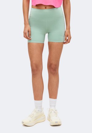 Vrouw draagt lichtgroene biker shorts met hoge taille, witte sportsokken en off-white hardloopschoenen, staand tegen een effen achtergrond.