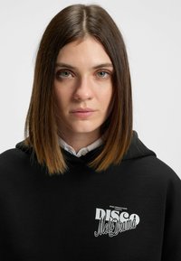 Felpa nera con cappuccio e cordoncino, caratterizzata da una grafica bianca con la scritta "DISCO Melodrama". Tessuto morbido, vestibilità casual.