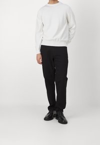 Weißer Strickpullover mit Rundhalsausschnitt und geripptem Saum, kombiniert mit schwarzen geraden Hosen und schwarzen Schnürschuhen. Einfaches Design, minimale Details.