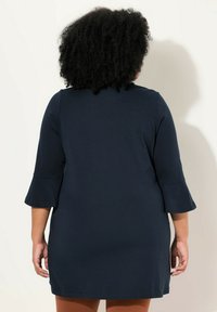 Mörkblå tunika med trekvarts ärmar och utsvängda ärmslut, tillverkad av ett mjukt tyg. Bakifrån visas en rak skärning och minimal detaljer.