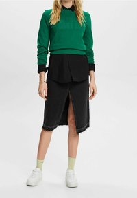 Sudadera verde con texto impreso, superpuesta a una camisa negra con botones. Falda de mezclilla negra con una abertura frontal y dobladillo deshilachado. Zapatillas blancas.