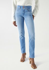 Vaqueros de mezclilla de color azul claro con una cintura alta, dos bolsillos frontales y un cierre de botón. Llevados con sandalias de tacón color beige y un suéter blanco texturizado.