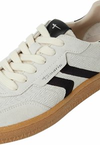 Hvite semskede sneakers med teksturdetaljer, svarte accenter og gummisåle. Har beige snørebånd og en sølvlogo på siden.