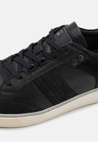 Svart läder- och mockaskor med texturerade paneler, runda snören, subtil varumärkeslogotyp och en lågt profilerad vit gummisula.