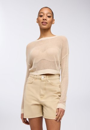 Maglione in maglia a rete di un beige chiaro, corto, abbinato a pantaloni denim a vita alta di un beige chiaro. Il maglione ha maniche lunghe e polsini a coste.