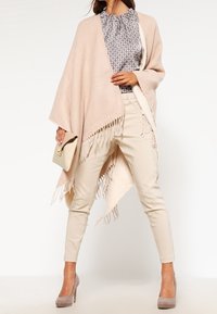 Poncho rosa chiaro con bordi frangiati, indossato sopra una camicetta a fantasia e pantaloni beige slim-fit, abbinato a tacchi taupe e una borsa a pochette.