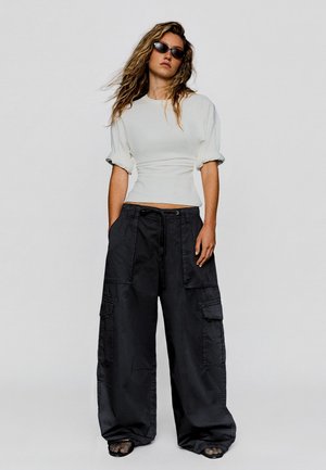 Femme portant un haut blanc cintré, un pantalon cargo noir oversize, des chaussures en résille noires et des lunettes de soleil noires, debout devant un fond uni.