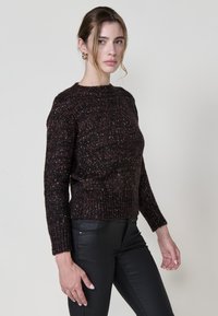 Dunkler, strukturierter Strickpullover mit rundem Halsausschnitt und langen Ärmeln, der dezente mehrfarbige Sprenkel aufweist. Kombiniert mit schwarzen, figurbetonten Hosen.