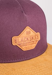 Casquette en tissu violet avec un patch en cuir marron portant le texte "BLACKSKIES" et un logo. Visière plate, motifs texturés sur le tissu.