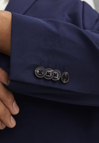 Ärmel einer marineblauen Anzugjacke mit glatter Textur, versehen mit vier schwarzen Knöpfen und kontrastierenden Nahtdetails. Hand, die den Bund greift.
