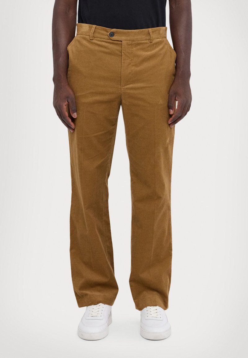 Pantalon en velours côtelé marron avec une coupe droite, doté de poches avant, d'une fermeture à un bouton et d'une surface texturée. Porté avec des baskets blanches.
