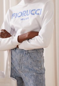 Sweatshirt blanc avec le logo bleu "FIORUCCI MILAN", associé à un jean en denim effet délavé clair orné d'un motif texte intégral.