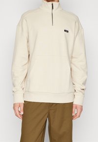 Sudadera de color crema con cremallera hasta un cuarto, confeccionada en un material suave, con un bolsillo delantero, cuello acanalado y un pequeño logo negro en el pecho.