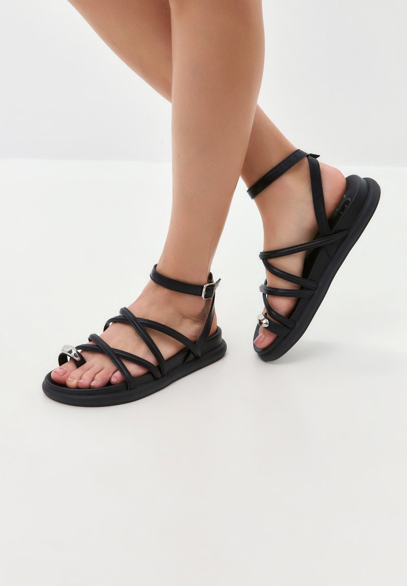 Sandalias negras de tiras hechas de goma con suela plana. Presentan múltiples bandas a lo largo del pie y un acento plateado brillante en la punta.