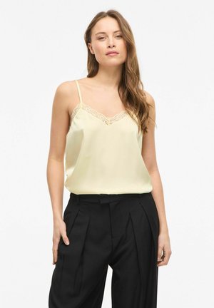 Jeune femme aux longs cheveux portant un caraco crème bordé de dentelle et un pantalon noir plissé taille haute, debout sur un fond blanc.