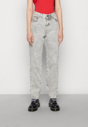 Personne portant un jean gris clair délavé à l'acide, des bottes noires à lacets et un top rouge sans manches, debout devant un fond blanc uni.