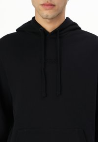 Hanorac negru realizat dintr-un material moale, cu glugă ajustabilă, logo-ul „GUESS” embossat și buzunar tip kangur la față.