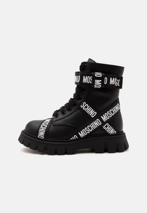 Black leather combat boot with thick sole, black laces, and white Moschino logo straps wrapped around ankle and foot.