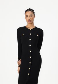 Robe tricotée noire à côtes avec manches longues, boutons dorés sur le devant et poches poitrine ; présente une fente sur le côté.