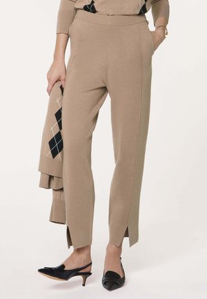 Pantalon en maille beige avec fentes frontales, porté avec des talons noirs slingback, tenant un pull assorti beige à motif losanges.