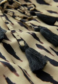 Tissu beige avec un imprimé animal noir présente un cordon tressé avec deux glands noirs, enrichissant la texture et le détail du design.