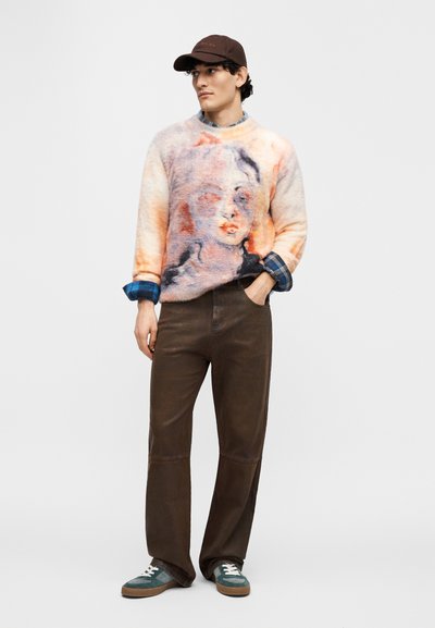 Πλεκτή μάλλινη ζακέτα με πολύχρωμο αφηρημένο σχέδιο προσώπου, με γυρισμένα μανίκια, συνδυασμένη με καφέ παντελόνι και τυρκουάζ αθλητικά παπούτσια. Καφέ καπέλο ολοκληρώνει το ντύσιμο.