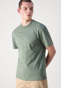 T-shirt en coton vert avec un col ras du cou, des manches courtes et une coupe ajustée. Présente un logo discret sur la poitrine et une texture lisse.