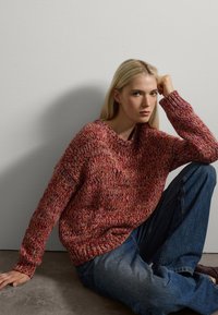 Suéter de punto en tonos rojos y naranjas con un corte holgado, puños acanalados y un escote amplio. Combinado con jeans de mezclilla azul.