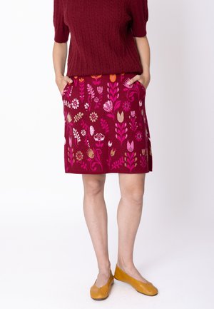 Donna che indossa un maglione rosso a maniche corte con trecce, gonna bordeaux a fantasia floreale con tasche e scarpe piatte giallo senape, in piedi.