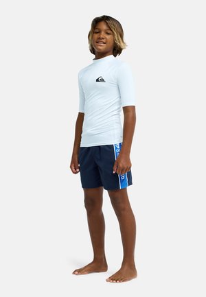 Jongen staat op blote voeten, draagt een wit rashguard met korte mouwen en marineblauwe zwembroek met een blauwe zijstreep.