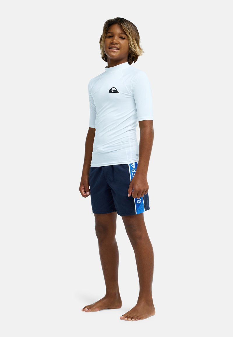 Chlapec stojaci naboso, oblečený v bielom rash garde s krátkym rukávom a námornícky modrých plavkách s modrým pruhom na boku.