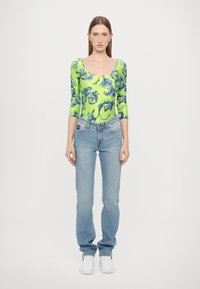 Langermet bodysuit i neon grønn med blå blomster mønster, sammen med lyseblå rette jeans og hvite sneakers.