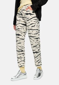 Beige Hose mit einem schwarzen Tigerstreifenmuster, ausgestattet mit hoher Taille, geraden Beinen und seitlichen Taschen, kombiniert mit silbernen Turnschuhen.